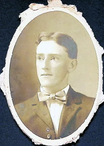 Portrait of a young man — Oval-vignette sepia studio portrait of a young man with combed-back hair wearing a suit jacket and a light bow tie. The print has been trimmed to an oval and its paper backing is torn and ragged around the edges. No text visible.
