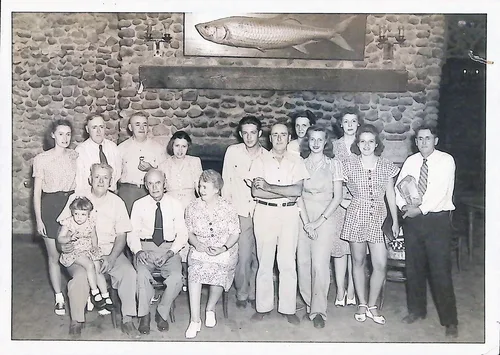 Group portrait at lodge — Black-and-white group portrait of about sixteen people, some seated and some standing, in front of a stone fireplace wall with a large mounted tarpon (fish) above them. A small child sits on a seated man's lap at left. No text visible.