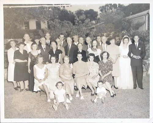 Large family gathering with bride — Black-and-white photo of a large group of roughly thirty adults and two small children posed outdoors in front of a house and garden. A bride in a white gown and veil stands at right beside a man in a dark suit, suggesting a wedding gathering. No text visible.