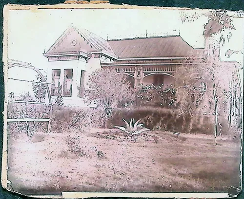 House with garden — Faded, pink-toned photograph of a single-story house with a hipped roof, a square bay/tower section with windows, and a covered porch with decorative trim. The overgrown yard has shrubs, an agave plant, and an arbor or trellis at left. No text visible.