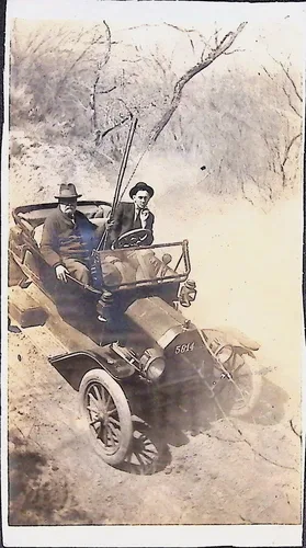 Two men in early automobile — Two men in hats and suits seated in an early open touring automobile on a dirt road or wash with bare trees behind. A fishing rod is propped up in the car. The license or number on the car reads "5814[?]". No handwritten text visible.