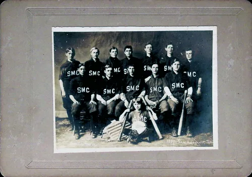 SMC baseball team — Studio team portrait of thirteen young men in dark uniforms lettered "SMC", arranged in rows with baseball bats, a glove, and a young mascot boy seated in front holding a bat. Mounted on a gray card. Faint imprint at lower right reads "...SAN ANGELO, TEX.[?]".