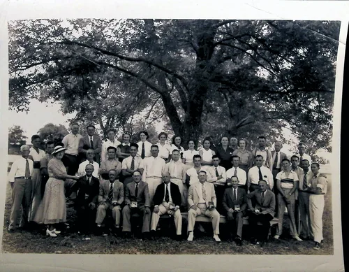 Large group under tree — Black-and-white photo of a large group of mostly men in shirtsleeves, ties, and suits, with a few women, posed in rows beneath a large spreading tree at what appears to be an outdoor gathering or reunion. Several front-row men hold cups or mugs. No text visible.
