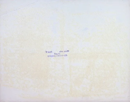 Blank verso with photo stamp — Largely blank cream-colored verso of a photograph. A faint blue ink stamp in the center reads, in part, "If mail [?] give credit line to [?] PHOTO CO." No handwritten identification.