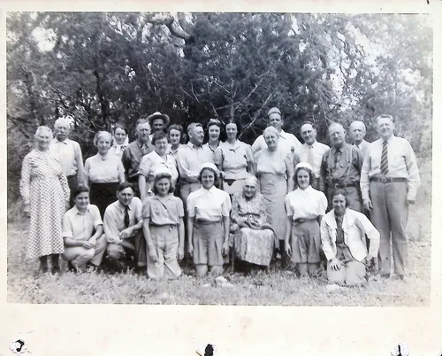 Menger Family group portrait — Outdoor black-and-white group portrait of about two dozen adults arranged in rows on grass in front of trees, including one elderly seated woman with a cane. (The verso, p015, identifies this as the "Menger Family" / H&H Company picnic.)