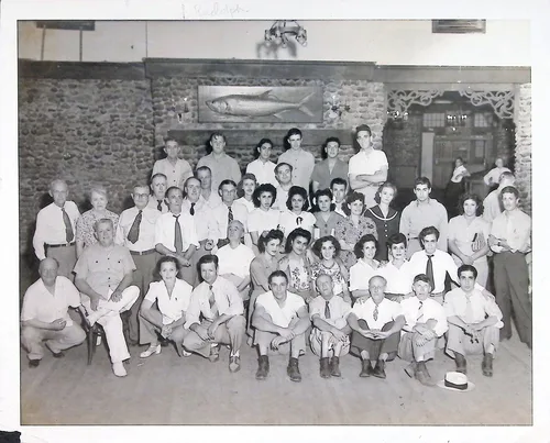 Large group portrait under fish — Black-and-white indoor group portrait of a large gathering (roughly forty people) posed in rows before a stone wall, with a mounted fish (tarpon) on a plaque above them. Faint pencil writing along the top edge is illegible.
