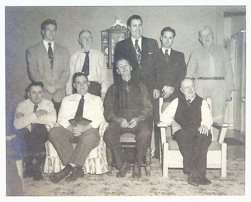 Group of nine men, studio portrait — Black-and-white indoor portrait of nine men in suits and ties, five standing and four seated in armchairs, in a furnished room with a wall clock cabinet. No visible text.