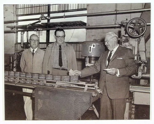 Three men at factory machinery — Black-and-white photo of three men beside industrial machinery in a factory; a conveyor line in front of them carries packages repeatedly labeled "Master Chef". One man at right appears to be inspecting a product.