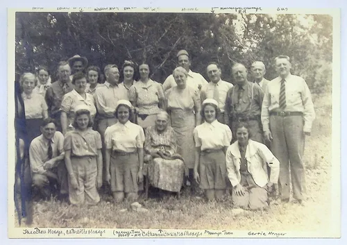 Menger family outdoor group — Sepia outdoor group portrait of about twenty people in rows before trees, with an elderly woman seated at center. Faint handwriting along the top edge is mostly illegible; ink captions along the bottom include "Theodore Menge[r], Catherine Menge[r]", "[?] Catharine [?] Barbara [?] Menger", "Mary[?]s Theo[?]", and "Gertie Menger".