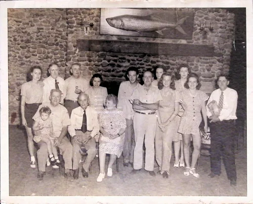 Family group under mounted fish — Black-and-white indoor group portrait of about fifteen people, including a small child on a man's lap, posed before a large stone fireplace with a mounted fish (tarpon) above. No clearly legible text.