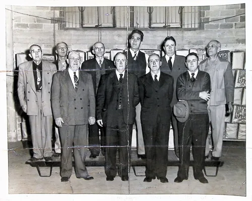 Group of ten men in warehouse — Black-and-white portrait of ten men in suits standing in two rows inside a warehouse; stacked crates and boxes behind them carry partly visible "COFFEE" labels. The print is heavily creased. Faint pencil writing along the bottom is illegible.