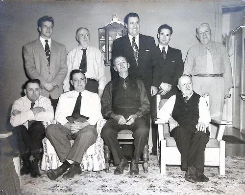 Group of nine men, studio portrait — Black-and-white indoor portrait of nine men in suits and ties, five standing and four seated in armchairs, in a furnished room with a wall clock cabinet (a near-duplicate framing of p018). No visible text.