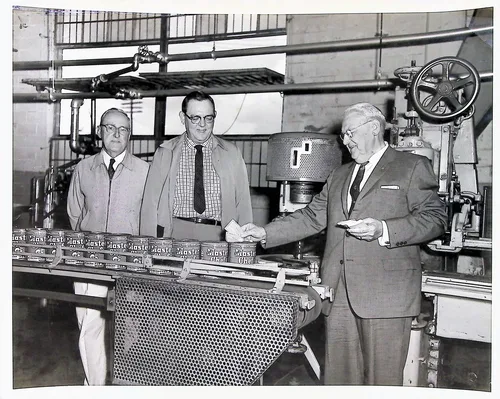 Three men at canning line — Black-and-white photo of three older men in suits standing at an industrial conveyor line where cans labeled "Master [?]" move along a perforated metal belt, in a factory or warehouse setting.