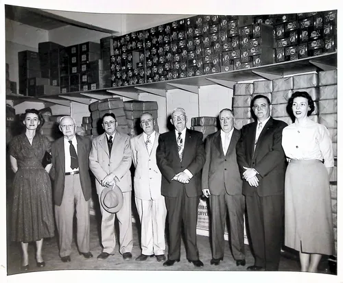 Group portrait in coffee warehouse — Black-and-white group portrait of seven men and two women standing in a row before stacks of boxed goods in a warehouse; a partial sign reading "...COFFEE" is visible behind them.