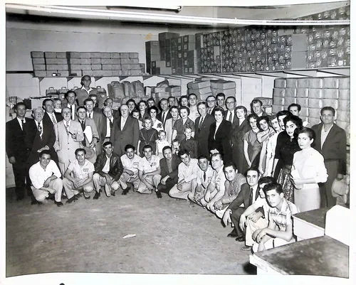 Large group portrait in warehouse — Black-and-white photo of a large group of roughly forty people, posed in standing and crouching rows inside a warehouse stacked with boxes. Several younger men in front wear matching work uniforms.