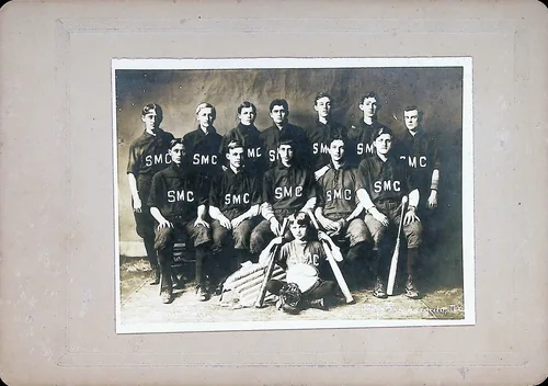 SMC baseball team portrait — Studio group portrait of a baseball/sports team of thirteen young men wearing dark jerseys lettered "SMC", with bats and a glove arranged in front. Mounted on gray card; a photographer's imprint reads "...ston, T[?]" at lower right.