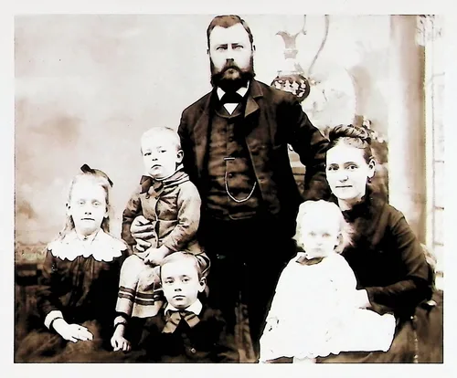 Bearded man with family — Studio family portrait of a bearded man standing center with a woman seated at right and four children, including a baby and a toddler in white. No text visible.