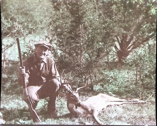 Hunter with deer (photograph) — Black-and-white photograph of a man in a cap and jacket kneeling in the woods, holding a rifle and posing beside a dead buck (deer with antlers). No text visible on the front.