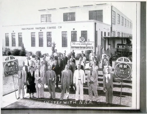 Hoffmann-Hayman Coffee Co. group — Large group of employees posed on steps outside a building marked "HOFFMANN HAYMAN COFFEE CO" with a banner reading "We are satisfied / HOFFMANN HAYMAN COFFEE Co" and two round "BUY NOW / H-H / QUALITY" signs. Caption along the bottom edge reads "IN STEP WITH N.R.A."