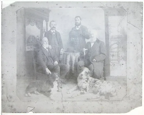 Group portrait with two dogs — Faded studio portrait of four men in an interior setting; three are seated and one stands at center with a beard. Two dogs lie on the floor in front. No visible text.