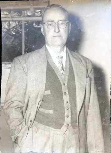 Portrait of an older man — Photo of an elderly man with glasses wearing a three-piece tweed suit, hands in pockets, standing before a window. No visible text.