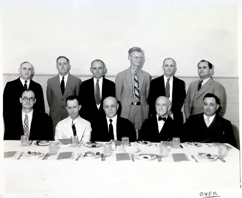 Group portrait at banquet table — Black-and-white photograph of eleven men, six standing and five seated, behind a set banquet table with plates and glasses. Handwritten "OVER" appears in the lower right corner, indicating a verso with identifications.