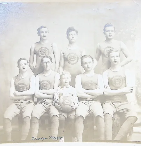 Basketball team portrait — Faded black-and-white team photograph of eight young men in athletic jerseys bearing a circular emblem, arranged in two rows, with a young boy seated in front holding a basketball. A faint ink signature appears at the bottom, possibly "[?] Menger".