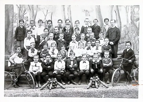 Group of boys with bicycles — Black-and-white group photograph of a large group of boys posed outdoors before a painted backdrop, with two bicycles at the sides and bowling pins arranged on the ground in front. An adult man stands at right rear. No legible text.