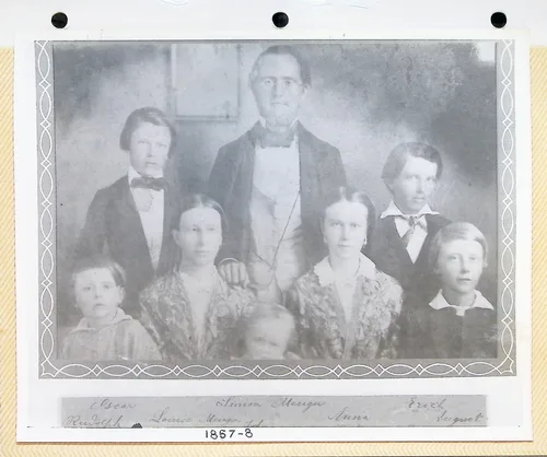 Menger family portrait, labeled — 1857-8 — Another print of the eight-person family group portrait (mounted on a punched album page), with handwritten names below the image: "Oscar", "Simon Menger", "Erich", "Rudolph", "Louise [?]", "Anna", "August", and the date "1857-8".