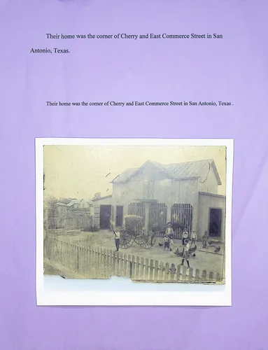 Home at Cherry & East Commerce St. — A captioned photograph mounted on purple paper. The typed caption (repeated) reads "Their home was the corner of Cherry and East Commerce Street in San Antonio, Texas." The old photo shows a two-story building/house with outbuildings, a picket fence, a horse-drawn buggy, and several people standing in the yard.
