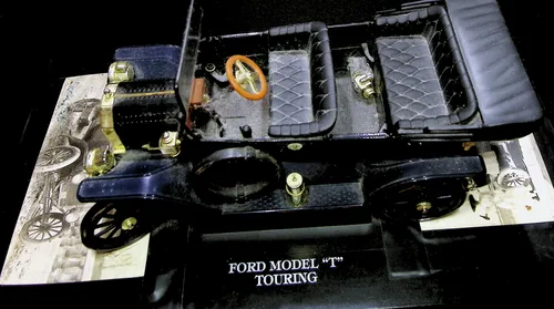 Ford Model T Touring (side view) — Color photo of a dark blue Ford Model T model car displayed at an angle, captioned "FORD MODEL \"T\" TOURING" on its base. An old sepia photo of a similar period car sits beneath it.