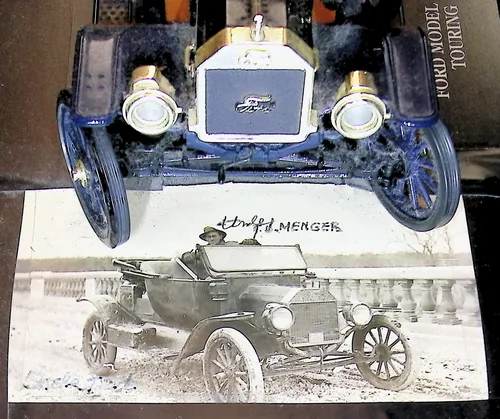 Model T photo signed Menger — An old black-and-white photo of a man seated in an early automobile (Model T) on a road by a balustrade, overlapping a color photo of the front of the blue model car. The photo is inscribed across the top "[?]rudof J. MENGER" with faint writing at the lower left.