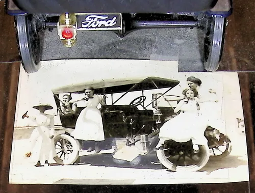 Women and children with Ford car — An old black-and-white photo of two women in long white dresses and two small children posed beside an early open automobile outdoors, overlapping a color photo of a model car's rear bumper bearing a "Ford" badge.